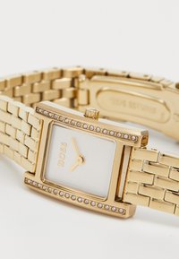 Montre rectangulaire dorée avec un cadran blanc, des accents en cristal et un bracelet lié. Comprend une couronne et le nom de la marque "BOSS" à 12 heures.