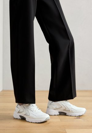 Chaussures de sport blanches avec une finition texturée, des lacets et des accents gris. Associées à un pantalon noir ample, sur un sol en bois clair.
