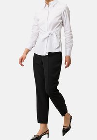 Biała koszula z długim rękawem z wiązaniem w talii, w zestawieniu z czarnymi, dopasowanymi spodniami i czarnymi szpilkami z wąskim czubkiem.