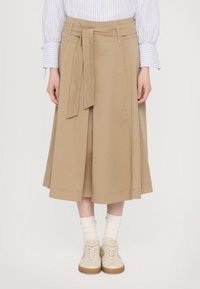 Vartalon alhaalta otettu kuva henkilöstä, joka on pukeutunut beige midi-hameeseen, jossa on solmittava vyö, valkoisiin sukkiin, vaalean kremi-kengissä ja raidallisen pitkähihainen paidan hihat näkyvät.