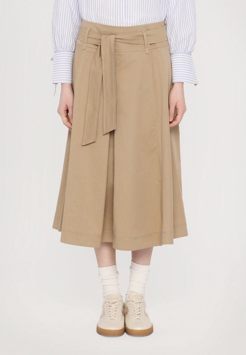 Vartalon alhaalta otettu kuva henkilöstä, joka on pukeutunut beige midi-hameeseen, jossa on solmittava vyö, valkoisiin sukkiin, vaalean kremi-kengissä ja raidallisen pitkähihainen paidan hihat näkyvät.