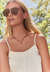 Brown mirrored sunglasses with metal wire frame, worn with a white, textured, spaghetti-strapped top and a gold chain necklace featuring a pendant.