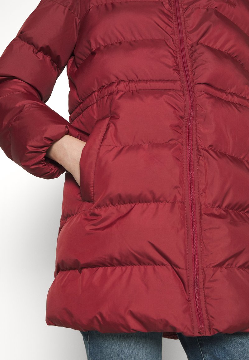 Persona che indossa un giubbotto trapuntato rosso con cerniera frontale, tasche laterali e mano infilata nella tasca sinistra, abbinato a jeans blu.