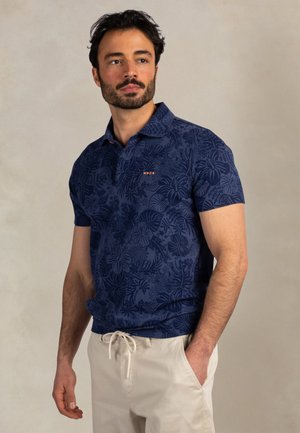 Man met baard, gekleed in een marineblauw poloshirt met bloemenprint en beige broek met trekkoord, staat met zijn rechterhand in de zak en kijkt naar links.
