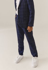 Tumesiniste ruudulise ülikonna tekstuuriga kangas, slim fit, koos valgete tossudega. Ülikond sisaldab ühe nupuga sulgemist ja tasku detaile.