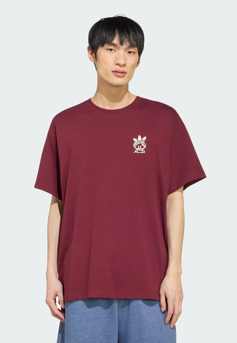 Junger Mann trägt ein lockeres dunkelrotes T-Shirt mit kleinem weißen Adidas- und Cartoon-Logo auf der Brust, kombiniert mit blauen Hosen.