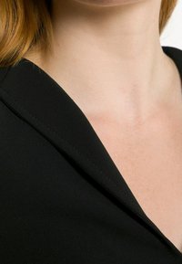 Czarna tkanina o gładkiej fakturze, z dekoltem w szpic. Zbliżenie podkreśla staranne szwy wzdłuż dekoltu.