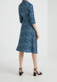 Kobieta ubrana w niebieską kwiecistą sukienkę do kolan z rękawami do trzech czwartej oraz czarnymi sandałami na obcasie, stojąca na gładkiej białej podłodze.