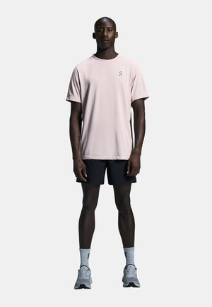Light pink short-sleeve t-shirt with crew neck and logo, paired with black shorts, gray sneakers, and gray socks in a neutral background.