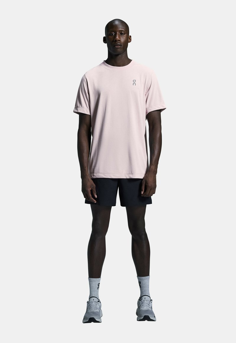 Lyserød kortærmet t-shirt med rund hals og logo, sammen med sorte shorts, grå sneakers og grå sokker på en neutral baggrund.