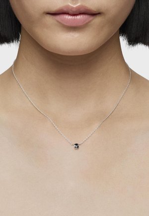 Collar de cadena de plata que presenta un pequeño charm de esmalte negro en forma de corazón, colocado sobre un cuello liso y desnudo.