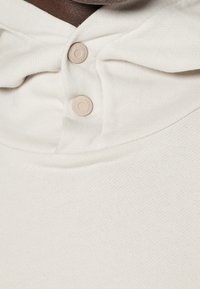 Felpa con cappuccio beige chiaro dalla texture morbida, con due bottoni rotondi di colore neutro e collo pieghevole.