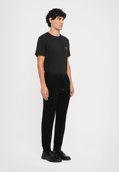 Fekete, rövid ujjú garbónyakú póló, bal mellrészen kis nyomott logóval, fekete, szűkített pizsamanadrággal és fekete cipővel párosítva.