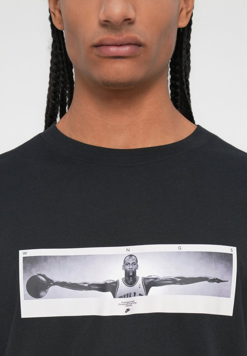 Schwarz Baumwoll-T-Shirt mit einem Grafikdruck eines Basketballspielers mit ausgestreckten Armen. Text und Logo unter dem Bild. Weiche Textur.
