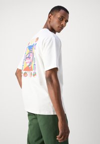 Camiseta de algodón blanca con un diseño gráfico colorido en la parte posterior, con patrones audaces y un corte relajado, combinada con pantalones verdes.