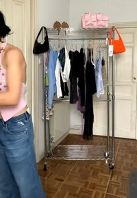 Kleiderständer mit verschiedenen hängenden Oberteilen, zwei Handtaschen an den Enden, High Heels und eine rosa Tasche auf dem oberen Regal, Frau in Jeans und pinkem Oberteil teilweise sichtbar.