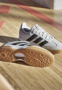 Weiße und schwarze Adidas Sneaker mit Gummisohlen auf einem Holzboden, im Hintergrund sind rote Spielfeldlinien sichtbar.