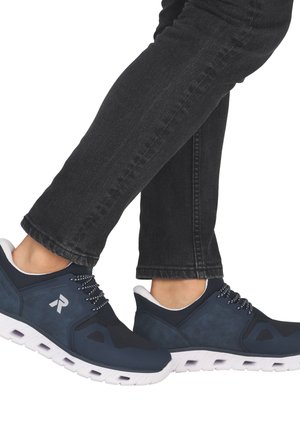 Personne portant un jean noir et des baskets de sport bleu marine avec des semelles blanches, debout avec un pied croisé sur l'autre.