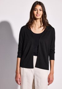 Cardigan nero con maniche lunghe, apertura frontale, texture morbida, indossato sopra un top nero e abbinato a pantaloni beige, caratterizzato da una vestibilità comoda.