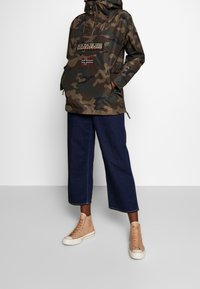 Kurtka anorak w wzór kamuflażu z przednią kieszenią, ozdobiona naszywką z logo i norweską flagą. Stylizowana z szerokimi dżinsowymi spodniami i beżowymi trampkami.