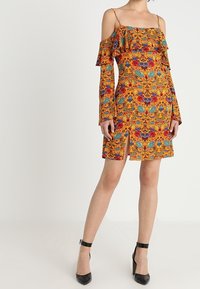 Vestido off-the-shoulder en un vibrante color naranja con patrones florales, volantes en el escote y las mangas, de longitud hasta la rodilla con una abertura lateral, combinado con tacones negros.