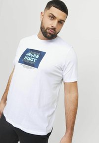 Valge puuvillane T-särk, millel on sinine graafiline ristkülikukujuline trükk, millel on tekst "JACK & JONES" ja horisontaalsed jooned. Lühikesed varrukad, mugav lõige.