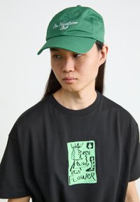 Casquette en coton vert avec "En Vacances" brodé en blanc ; portée avec un t-shirt noir arborant un grand motif graphique vert.