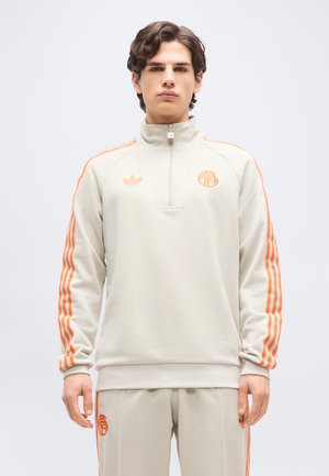 Joven vestido con un chándal beige claro con franjas y logotipos naranjas, de pie frente a un fondo blanco liso.