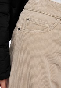 Beige corduroy pants with vertical ribbing, featuring a side pocket and a small branded logo. The material appears soft and textured.