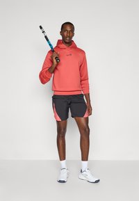 Un sweat à capuche corail avec un logo sur le devant, un short noir avec des accents roses, des chaussettes blanches et des chaussures de sport, tenant une raquette de tennis.