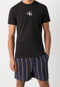 Svart bomullst-shirt med en flerfärgad "Calvin Klein Jeans" logga, parat med marinblå randiga shorts med röda och vita detaljer.