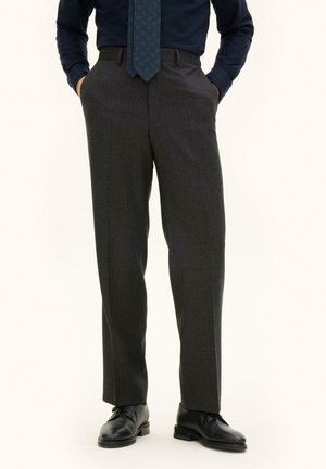 Uomo che indossa pantaloni da abito grigio scuro, scarpe eleganti nere, camicia blu navy e cravatta blu scuro a fantasia, in piedi con le mani nelle tasche.