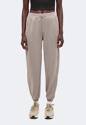 Personne portant un pantalon de jogging beige avec taille à cordon, poignets élastiqués, chaussettes blanches, baskets beiges et un haut court noir.