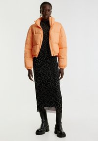 Gekürzte, voluminöse orangefarbene Jacke mit hohem Kragen, getragen über einem schwarzen Midikleid mit Punkten. Modell trägt schwarze klobige Stiefel.