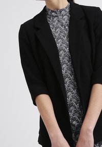 Černý blazer s strukturovaným designem, se třičtvrtě rukávy, nošený přes vzorovaný černobílý top s vysokým límcem.