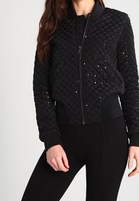 Czarna pikowana kurtka bombowiec z zamkiem z przodu, teksturowaną powierzchnią, prążkowanymi mankietami i rozrzuconymi cekinami jako ozdoba. W zestawie z czarnymi legginsami.