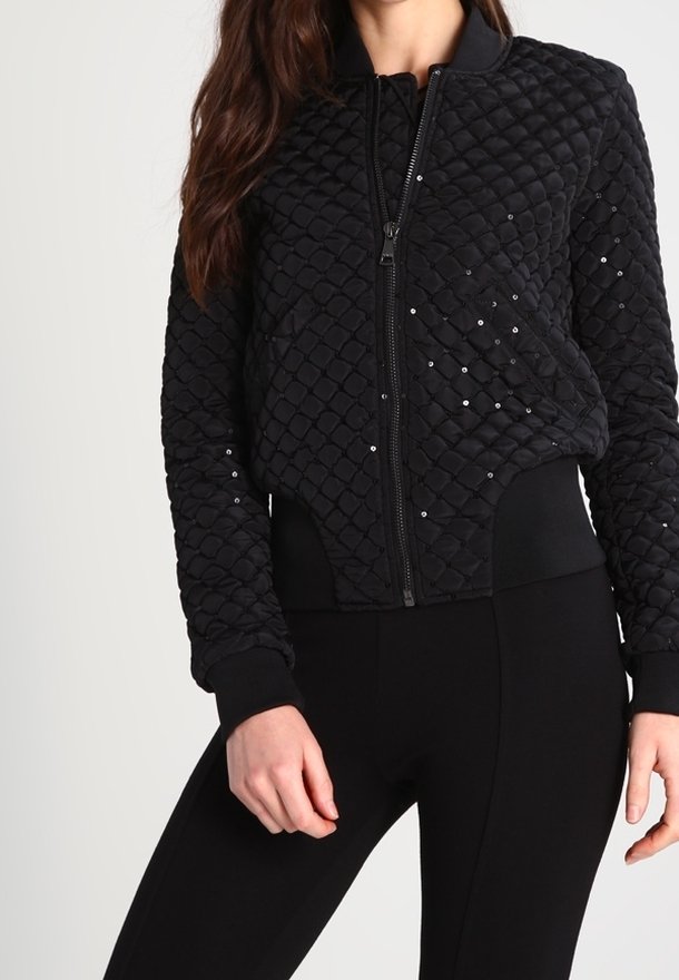 Czarna pikowana kurtka bombowiec z zamkiem z przodu, teksturowaną powierzchnią, prążkowanymi mankietami i rozrzuconymi cekinami jako ozdoba. W zestawie z czarnymi legginsami.