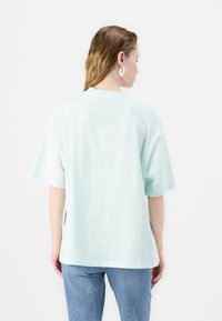Světle mákově zelené oversized tričko, krátké rukávy, bavlněný materiál, s vyraženým textem na zadní straně, nošeno s modrými d�žínami.