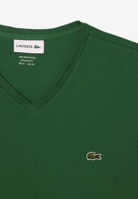 T-shirt verde a scollo a V realizzato in 100% cotone pima, con un piccolo logo di coccodrillo ricamato sul lato sinistro del petto, texture liscia e vestibilità regolare.