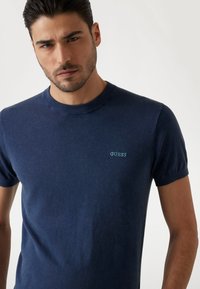 Camiseta de manga corta de color azul marino, hecha de una tela suave, con cuello redondo y un pequeño logo "GUESS" en turquesa en el área del pecho izquierdo.