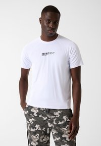 Weißes sportliches T-Shirt mit kurzen Ärmeln und Netzstruktur, mit einem reflektierenden "GUESS"-Logo auf der Brust. Kombiniert mit gemusterten dunklen Hosen.