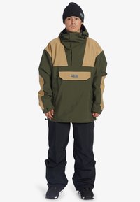 Chaqueta impermeable verde y marrón con capucha, bolsillo frontal y puños ajustables. Combinada con pantalones de nieve negros y botas negras.