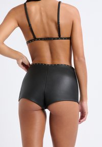 Top bikini in ecopelle nera con spalline regolabili, borchie dorate lungo la fascia, e pantaloni shorts a vita alta abbinati con una superficie liscia.