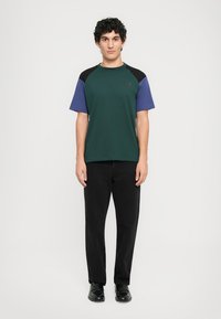 T-shirt verde a maniche corte con dettagli neri e viola, abbinato a pantaloni neri. Il tessuto appare liscio; la T-shirt ha un collo rotondo.