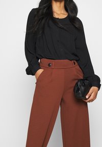 Černá košile se zapínáním na knoflíky, dlouhými rukávy a elastickými manžetami, ve spojení s hnědými kalhotami s vysokým pasem, předními kapsami a zapínáním na knoflíky.