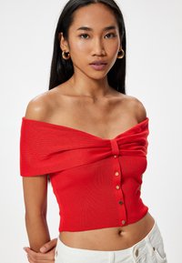 Rød off-shoulder top med sløjfedetail, ribbet tekstur og guldknap accenter; tætsiddende design kontrasterer med lyse bukser.