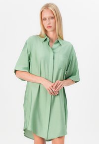 Rochie din bumbac cu manșon scurt, de culoare verde deschis, cu guler structurat, buzunar frontal, închidere cu nasturi pe față și fante laterale pentru ușurința mișcării. Textură netedă.