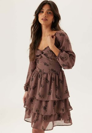 Kvinde med langt mørkt hår iført en lagdelt malvafarvet kjole med lange ærmer og sorte blomsterprint, poserer med hånden nær skulderen.