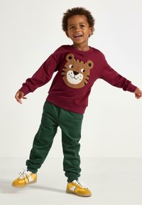 Mörkröd sweatshirt med en tigeransiktsdesign, kombinerad med gröna kordbyxor och gula sneakers, med texturerade material och kontrasterande färger.