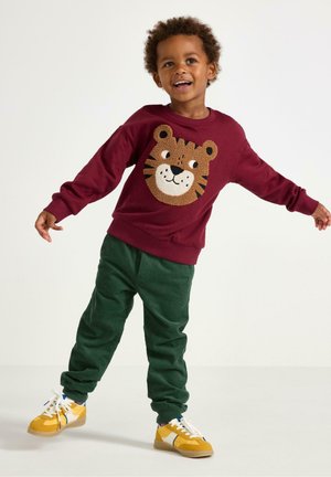 Mörkröd sweatshirt med en tigeransiktsdesign, kombinerad med gröna kordbyxor och gula sneakers, med texturerade material och kontrasterande färger.